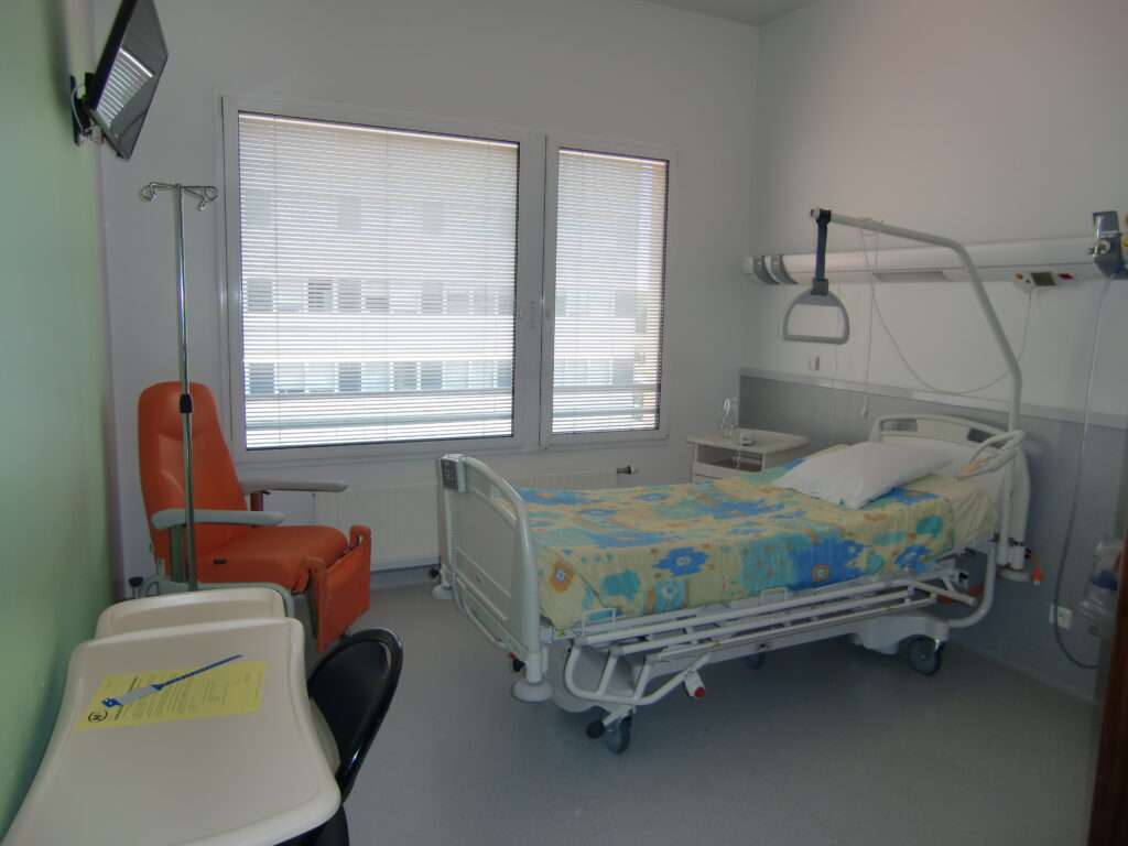 Chambre de l'hôpital avec le lit à droite, le fauteuil, la télé et une table amovible à gauche.